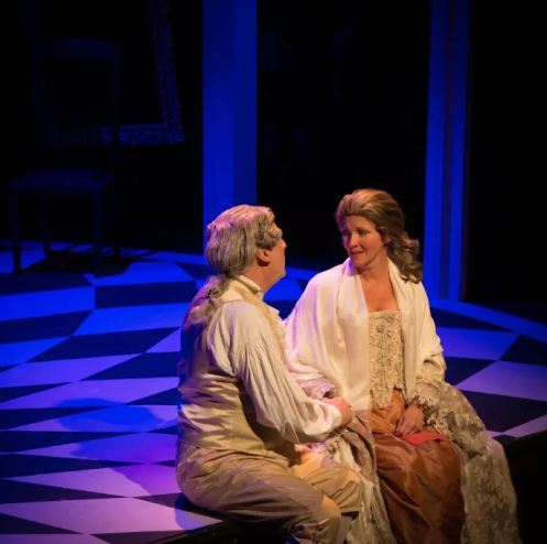 A man and woman sitting together on stage, engaged in conversation during a theatrical performance. The scene is illuminated with purple lighting, highlighting the checkerboard floor and the intimate atmosphere.