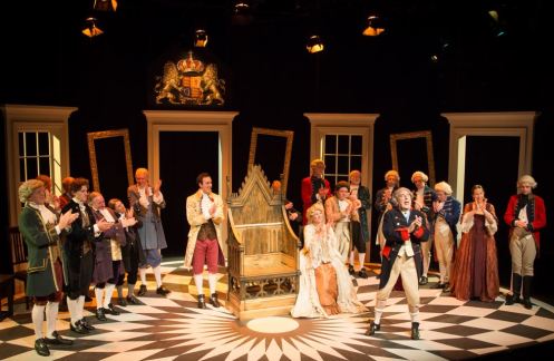 A theatrical scene featuring a group of actors in period costumes, gathered on a stage with a throne-like chair in the center. The actors are in a lively performance, some applauding, while others are engaged in the dialogue or action. The stage is decorated with curtains and characterized by a patterned floor.