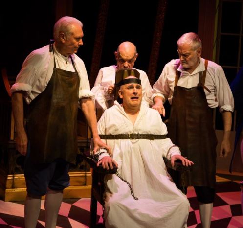 Four actors in historical costumes performing in a theatrical setting. One actor, seated and restrained in a chair, displays a distressed expression while others stand over him.