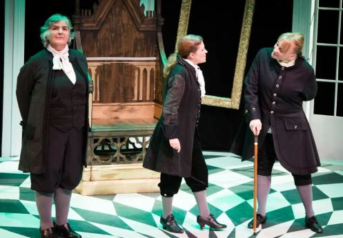 Three actors dressed in historical costumes on stage, performing in a theatrical production with a checkerboard floor and a stylized backdrop.