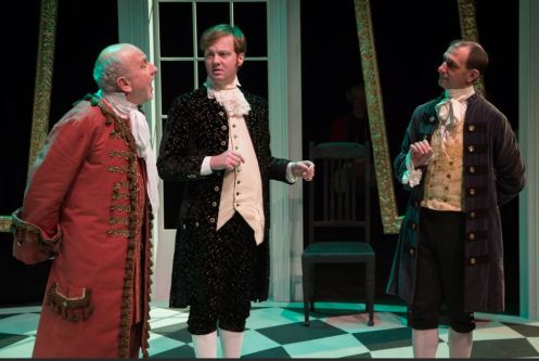 Three actors performing in period costumes during a theatrical scene, with a checkered floor and a backdrop featuring windows.